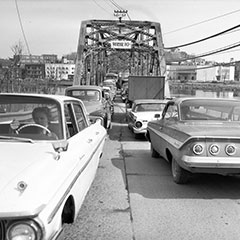 Bouchon de circulation sur un pont de fer où une dizaine de voitures se trouvent. À l'arrière-plan, nous pouvons voir une ville.
