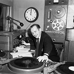 Animateur de radio derrière le micro. Il y a deux tables tournantes, le tableau de commande et autres composantes du studio.