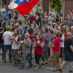 Protesters, dressed in red, are banging on pots and pans. The Québec flag and a red flag are floating above them.