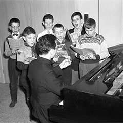Un petit groupe d'enfants pratique un chant choral auprès de l'abbé Claude Thompson assis au piano.