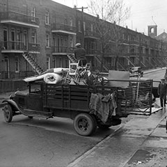 Scène de déménagement où un camion est rempli de meubles, de sommiers et de chaises. Un homme est monté sur le chargement.